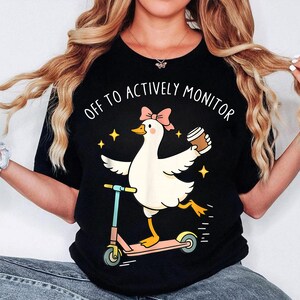 Op de afbeelding: Zwart T-shirt met een cartoon gans op een roze scooter, met een koffiekopje. De gans draagt een roze strik en de tekst luidt "OFF TO ACTIVELY MONITOR". Het ontwerp bevat ook gele sterren.