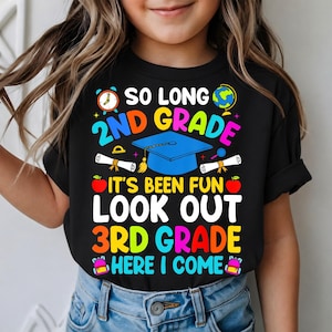May include: Black t-shirt with a colorful graphic celebrating the end of 2nd grade and the start of 3rd grade. The design includes a graduation cap, diplomas, an apple, and the text "So Long 2nd Grade, It's Been Fun, Look Out 3rd Grade, Here I Come."