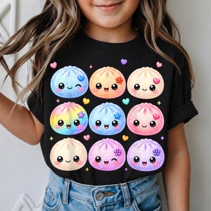 Könnte beinhalten: Ein schwarzes T-Shirt mit einem Grafikdruck von neun bunten, glitzernden, runden Teigtaschen mit lächelnden Gesichtern. Jede Teigtasche hat eine einzigartige Pastellfarbe, darunter Blau, Orange, Pink und Regenbogen, verziert mit kleinen Herzen und Blumen.