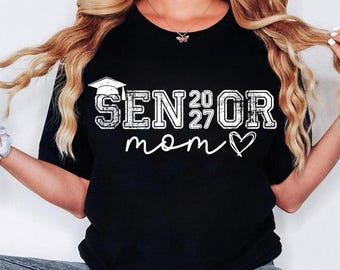 May include: Black t-shirt with the text "SENIOR 2027 mom" in white, featuring a graduation cap graphic. The design includes a heart symbol. The shirt is a casual style.