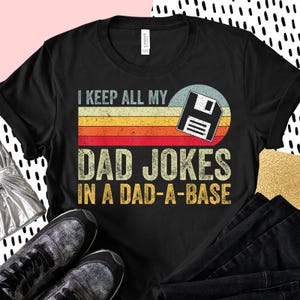Op de afbeelding: Zwart T-shirt met de tekst "I KEEP ALL MY DAD JOKES IN A DAD-A-BASE" in witte en gele letters. Het ontwerp bevat een retro zonsondergangafbeelding met een floppy disk-pictogram. Het shirt wordt gecombineerd met een zwarte broek en sneakers.