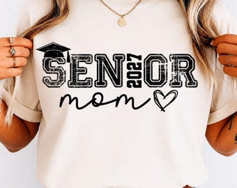 May include: A cream-coloured t-shirt featuring the text "SENIOR 2027 mom" in black, with a graduation cap above "SENIOR" and a heart symbol. The shirt has a relaxed fit.