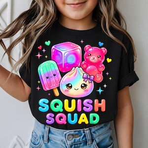 May include: A black t-shirt featuring a colorful graphic of a pink gummy bear, a gradient popsicle, a glowing cube, and a smiling dumpling with a purple bow. The text SQUISH SQUAD is printed in bright, multi-colored bubble letters below the icons.