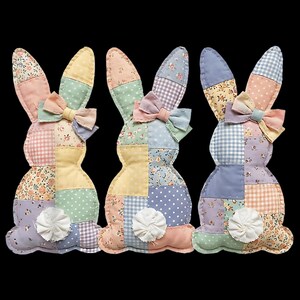 Puede incluir: Tres figuras decorativas de conejos de Pascua. Cada conejo está hecho de tela de patchwork en colores pastel, incluyendo rosa, amarillo, azul y verde. Tienen pajaritas y colas blancas y rizadas. Los conejos están colocados sobre un fondo negro.