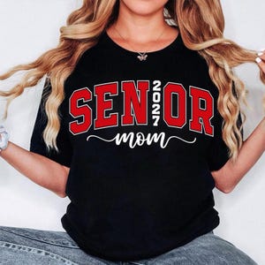 K&ouml;nnte beinhalten: Schwarzes T-Shirt mit dem Aufdruck "SENIOR 2027 mom" in Rot und Wei&szlig;. Das Wort "SENIOR" ist in fetten Blockbuchstaben mit roter Umrandung gehalten. Das Jahr "2027" ist vertikal zwischen "SENIOR" und "OR" gestapelt. Das Wort "mom" ist kursiv geschrieben.