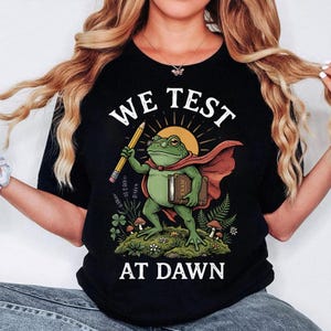 Op de afbeelding: Zwart T-shirt met een afbeelding van een kikker met een cape en een potlood en een boek. De tekst "WE TEST AT DAWN" staat boven en onder de kikkerillustratie. De kikker is groen met een bril en een rode cape.