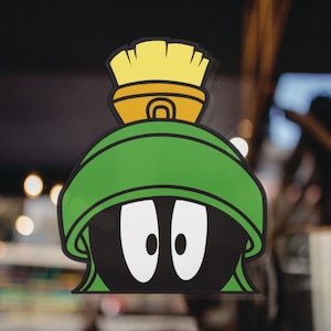 Puede incluir: Una pegatina con el personaje de dibujos animados Marvin el Marciano. La pegatina muestra la cabeza de Marvin con un casco verde, ojos blancos grandes y un objeto en forma de cepillo amarillo y naranja en la parte superior. El fondo est&aacute; borroso.
