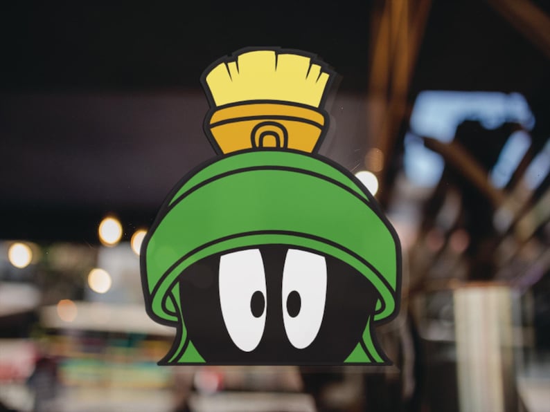Puede incluir: Una pegatina con el personaje de dibujos animados Marvin el Marciano. La pegatina muestra la cabeza de Marvin con un casco verde, ojos blancos grandes y un objeto en forma de cepillo amarillo y naranja en la parte superior. El fondo est&aacute; borroso.