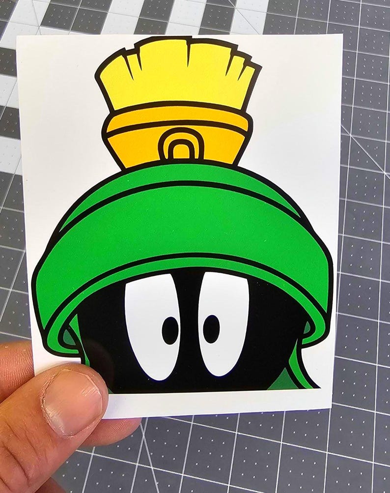 Puede incluir: Una pegatina con el personaje de dibujos animados Marvin el Marciano. La pegatina muestra la cabeza negra de Marvin, sus grandes ojos blancos, su casco verde y su tocado amarillo y naranja. La pegatina est&aacute; sobre un fondo blanco.
