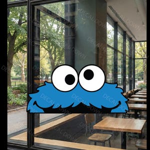 May include: A blue, furry-textured character decal featuring two large, white, googly eyes with black pupils, positioned on a glass window. The design includes a shaggy blue top edge and a wide, black, open-mouthed expression at the bottom.