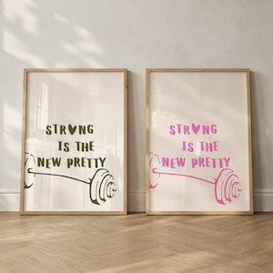 Pode incluir: Duas impressões emolduradas com o texto "STRONG IS THE NEW PRETTY". Uma impressão tem texto verde e uma ilustração de haltere verde, a outra tem texto rosa e uma ilustração de haltere rosa. Ambas estão em molduras de madeira clara.
