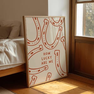 Könnte beinhalten: Ein rechteckiger gerahmter Kunstdruck mit weißem Hintergrund, einem sich wiederholenden Muster aus roten Hufeisenillustrationen und dem Text "HOW LUCKY ARE WE", der in roten Großbuchstaben in der Mitte abgebildet ist.