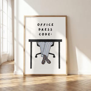 以下が含まれることがあります： 黒い文字で「OFFICE DRESS CODE:」と書かれた黒いデスクのフレーム入りアートプリント。デスクの下には、水色と白のストライプのズボンを履き、水色の水玉模様が入った茶色の靴下を履いた脚が椅子に座っている様子が描かれています。