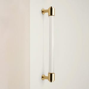 May include: A clear acrylic and brass door pull handle. The handle has a cylindrical acrylic center and brass mounting hardware. The brass hardware is a bright gold color. The handle is mounted on a white surface.