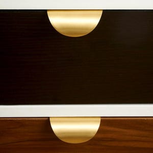 Può includere: Primo piano di un moderno comò a due cassetti. I cassetti sono marrone scuro e noce con una finitura bianca. Ogni cassetto ha una maniglia dorata a forma di semicerchio. Una sciarpa azzurra e un tulipano rosa sono sullo sfondo.