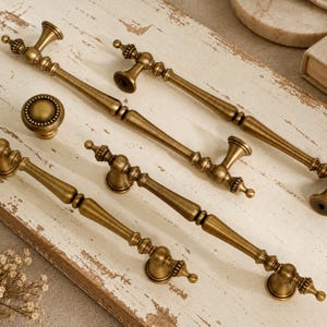 Op de afbeelding: Een verzameling antiek messing beslag, bestaande uit drie lange, slanke kastgrepen met decoratieve uiteinden en een ronde, met kralen versierde kastknop. Elk stuk heeft een geborstelde metalen afwerking met donkere, verdiepte details.