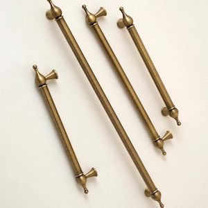May include: Four antique brass cabinet pulls arranged diagonally. Each handle features a long, cylindrical bar with a smooth finish, capped at both ends by decorative, rounded finials and flared, conical mounting posts for attachment.