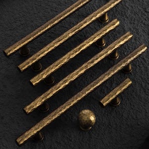 May include: A collection of five long, cylindrical cabinet pulls and one round cabinet knob, all featuring a dark bronze finish with a distinct hammered, textured surface and conical mounting posts for attachment to furniture or cabinetry.