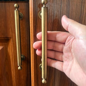 May include: A pair of cylindrical cabinet pulls with a brushed brass finish. Each handle features a long, straight central bar with a small spherical knob at both the top and bottom ends, mounted to the wood cabinet doors with circular bases.