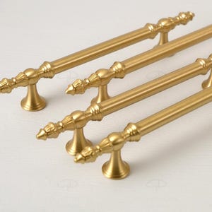 May include: Four gold-colored cabinet pulls with ornate detailing. Each pull features a long, straight bar with decorative ends and a central design. The pulls are mounted on small, flared bases, creating an elegant and classic look for furniture.