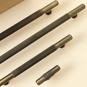 May include: Four antique bronze cabinet pulls with a ribbed design. The pulls are in varying lengths, with a small knob-style pull at the end. The hardware is set against a neutral background.