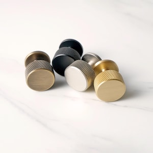 Puede incluir: Una colección de pomos de armario en varios acabados metálicos, incluyendo latón, negro y plata. Cada pomo presenta un agarre texturizado y moleteado y una cara plana y circular. Los pomos están dispuestos sobre una superficie de mármol blanco.