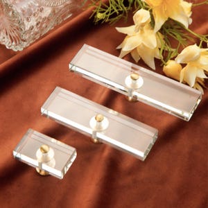 Op de afbeelding: Drie heldere rechthoekige kastknoppen met goudkleurige hardware. De knoppen zijn gemaakt van een helder, transparant materiaal en zijn gerangschikt op een bruin oppervlak. De kastknoppen zijn van verschillende grootte, met een kleine knop links en twee grotere knoppen rechts.