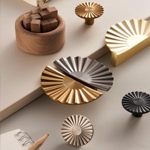 May include: An assortment of decorative hardware, including cabinet pulls and knobs. The hardware features a fan-like design in gold, silver, and black finishes. A wooden bowl with wooden blocks is also present.