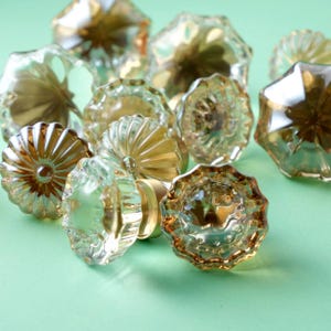 Könnte beinhalten: Eine Sammlung von Schrankknöpfen aus klarem und bernsteinfarbenem Glas mit verschiedenen facettierten und floralen Designs. Die Knöpfe sind auf einer hellgrünen Oberfläche angeordnet, die ihre reflektierenden Eigenschaften und komplizierten Details hervorhebt. Die Hardware hat eine messingfarbene Basis.