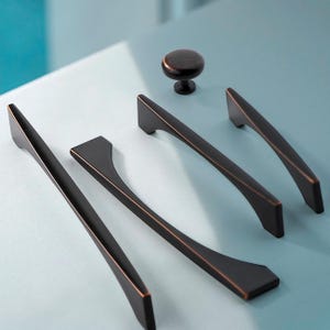 May include: A collection of bronze-colored cabinet hardware, including three long, curved handles and one round knob. The hardware is displayed on a light blue surface, showcasing the sleek, modern design.
