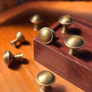 May include: A collection of round cabinet knobs with a brushed antique brass finish. Each knob features a smooth, domed top and a tapered base. Several knobs are arranged on a dark wood block, while others are scattered nearby on a wooden surface.