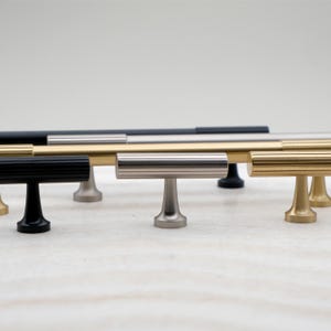 K&ouml;nnte beinhalten: Eine Sammlung von Schrankgriffen in verschiedenen metallischen Ausf&uuml;hrungen, darunter Schwarz, Gold, Silber und geb&uuml;rstetes Nickel. Die T-f&ouml;rmige Hardware verf&uuml;gt &uuml;ber eine horizontale Stange mit einem vertikalen Sockel, geeignet f&uuml;r Schubladen und Schrankt&uuml;ren.