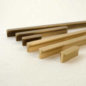 Könnte beinhalten: Eine Auswahl an Messing-Schrankgriffen in verschiedenen Größen. Die Griffe haben eine rechteckige Stangenform mit abgerundeten Kanten und einem warmen, metallischen Finish. Die Hardware ist diagonal angeordnet und zeigt die verschiedenen Längen und Farbtöne.