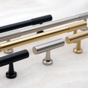 K&ouml;nnte beinhalten: Eine Auswahl an Schrankgriffen, darunter T-Bar- und Stangengriffe. Die Griffe sind in den Ausf&uuml;hrungen Mattschwarz, Nickel geb&uuml;rstet und Gold erh&auml;ltlich. Die T-Bar-Griffe haben ein zylindrisches Design mit horizontalen Rillen. Die Stangengriffe sind lang und schlank.