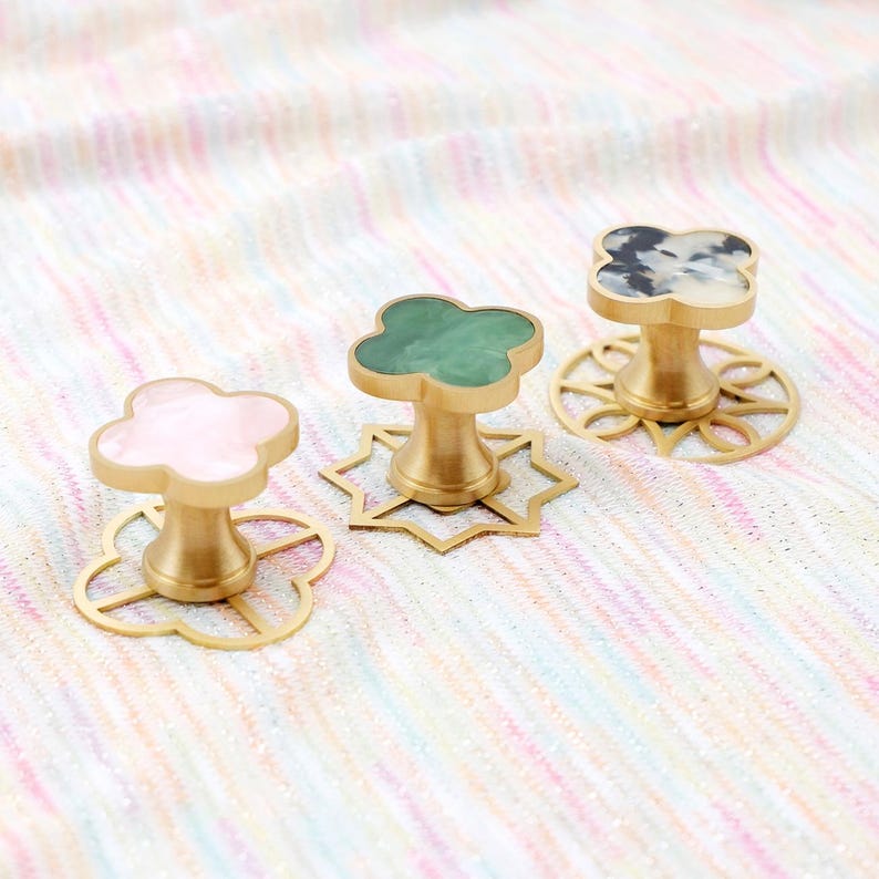 Op de afbeelding: Drie decoratieve kastknoppen met klavervormige toppen in roze, groene en zwart-witte patronen. De knoppen hebben een goudkleurige basis en staan op decoratieve goudkleurige bases. De knoppen staan op een veelkleurige stof.