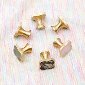 Op de afbeelding: Vijf messingkleurige kastknoppen met klavervormige bovenkanten. E&eacute;n knop heeft een roze oppervlak, een andere heeft een moza&iuml;ekachtig oppervlak en een derde heeft een groen oppervlak. De knoppen zijn gerangschikt op een veelkleurige stof.