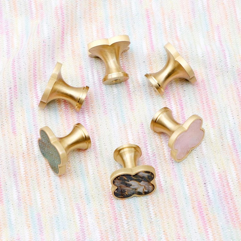 Op de afbeelding: Vijf messingkleurige kastknoppen met klavervormige bovenkanten. E&eacute;n knop heeft een roze oppervlak, een andere heeft een moza&iuml;ekachtig oppervlak en een derde heeft een groen oppervlak. De knoppen zijn gerangschikt op een veelkleurige stof.