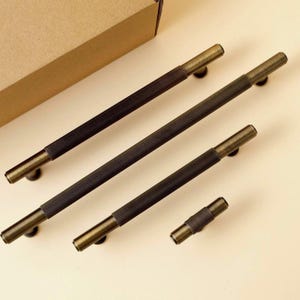 Pu&ograve; includere: Un set di quattro maniglie per armadietti di varie lunghezze, caratterizzate da una finitura in ottone antico scuro. Ogni maniglia presenta una sezione centrale con una fine texture a coste verticali ed estremit&agrave; cilindriche lisce.