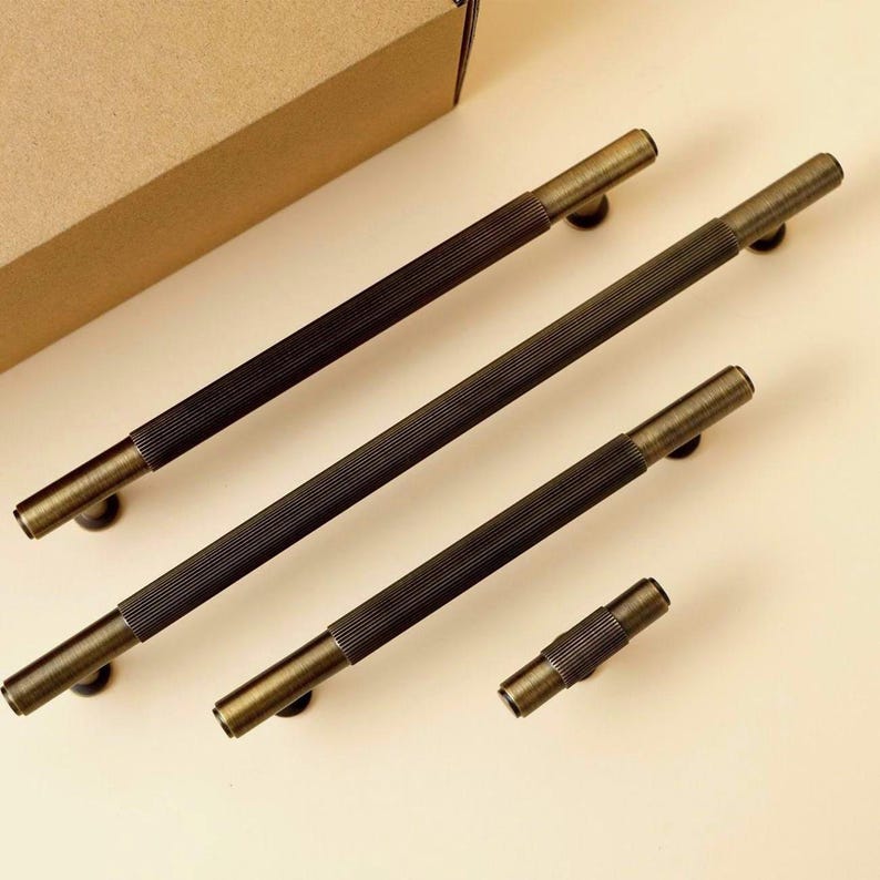 Pu&ograve; includere: Un set di quattro maniglie per armadietti di varie lunghezze, caratterizzate da una finitura in ottone antico scuro. Ogni maniglia presenta una sezione centrale con una fine texture a coste verticali ed estremit&agrave; cilindriche lisce.