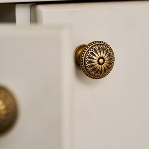 May include: Close-up of a decorative brass cabinet knob with a floral design. The knob is round with a detailed pattern and is mounted on a white cabinet door. The knob has an antique finish.