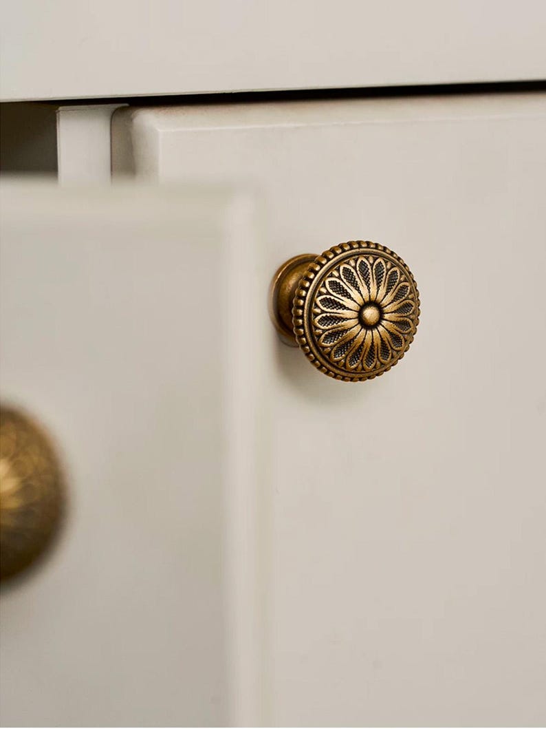 May include: Close-up of a decorative brass cabinet knob with a floral design. The knob is round with a detailed pattern and is mounted on a white cabinet door. The knob has an antique finish.