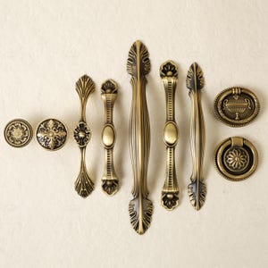 May include: An assortment of antique brass cabinet hardware, including knobs, pulls, and handles. The hardware features ornate floral and leaf designs, with a warm, aged finish. Various sizes and styles are displayed, suitable for furniture restoration or new projects.