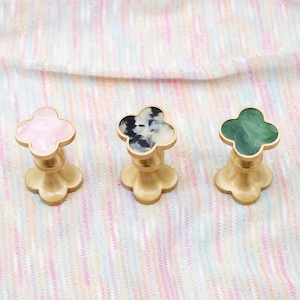 Op de afbeelding: Drie decoratieve kastknoppen met een klaverbladontwerp. De knoppen hebben een goudkleurige basis en een bovenkant in roze, zwart en wit en groen. De knoppen worden gepresenteerd op een veelkleurig, gestructureerd oppervlak.