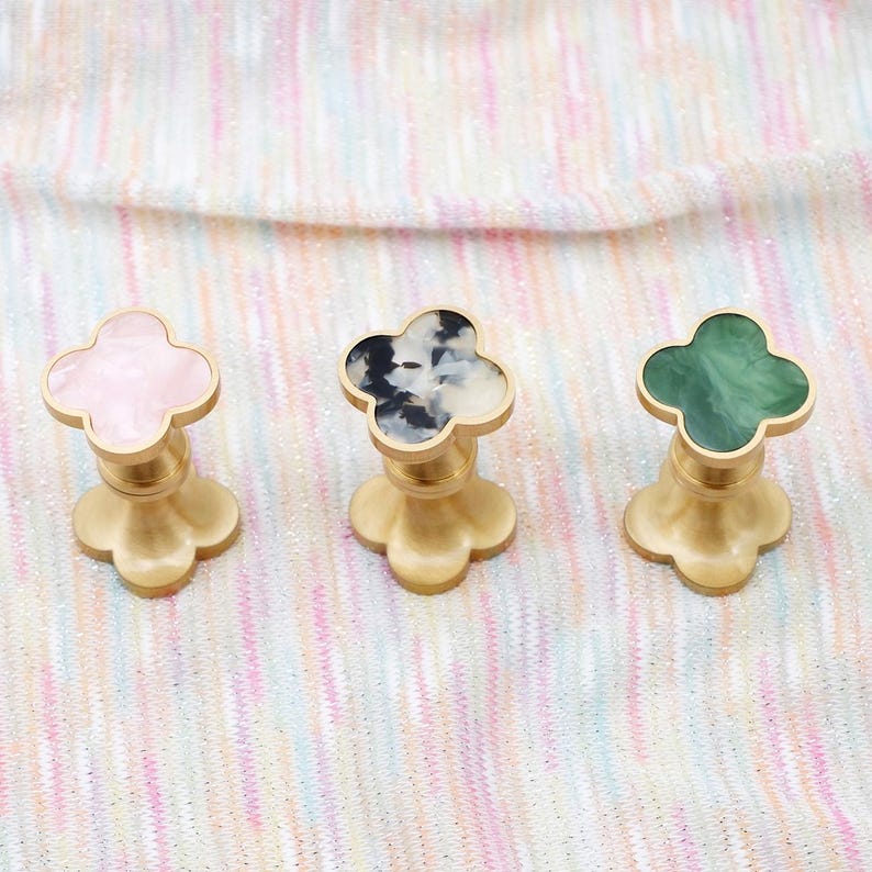 Op de afbeelding: Drie decoratieve kastknoppen met een klaverbladontwerp. De knoppen hebben een goudkleurige basis en een bovenkant in roze, zwart en wit en groen. De knoppen worden gepresenteerd op een veelkleurig, gestructureerd oppervlak.