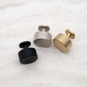 K&ouml;nnte beinhalten: Drei moderne Schrankkn&ouml;pfe in Schwarz, Silber und Gold. Jeder Knopf hat einen zylindrischen K&ouml;rper mit vertikalen Rillen und einer kreisf&ouml;rmigen Oberseite. Die Kn&ouml;pfe sind f&uuml;r eine einfache Installation konzipiert und verleihen jedem M&ouml;belst&uuml;ck Eleganz.