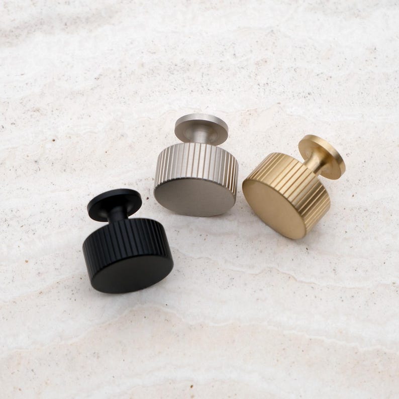 K&ouml;nnte beinhalten: Drei moderne Schrankkn&ouml;pfe in Schwarz, Silber und Gold. Jeder Knopf hat einen zylindrischen K&ouml;rper mit vertikalen Rillen und einer kreisf&ouml;rmigen Oberseite. Die Kn&ouml;pfe sind f&uuml;r eine einfache Installation konzipiert und verleihen jedem M&ouml;belst&uuml;ck Eleganz.
