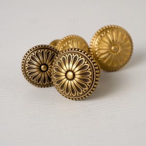 May include: Close-up of several antique brass cabinet knobs. Each knob features a detailed floral design with a central button and a beaded outer edge. The knobs are arranged on a white surface, showcasing their intricate details.