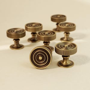May include: A collection of seven circular cabinet knobs made of antique brass-toned metal. Each knob features a raised center point surrounded by concentric circular ridges and a textured, ribbed outer edge, supported by a pedestal base.