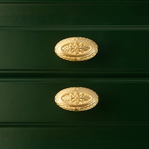 Puede incluir: Primer plano de dos tiradores de caj&oacute;n ovalados dorados con un dise&ntilde;o de estrella sobre un mueble verde oscuro. Los tiradores tienen una superficie detallada y texturizada.