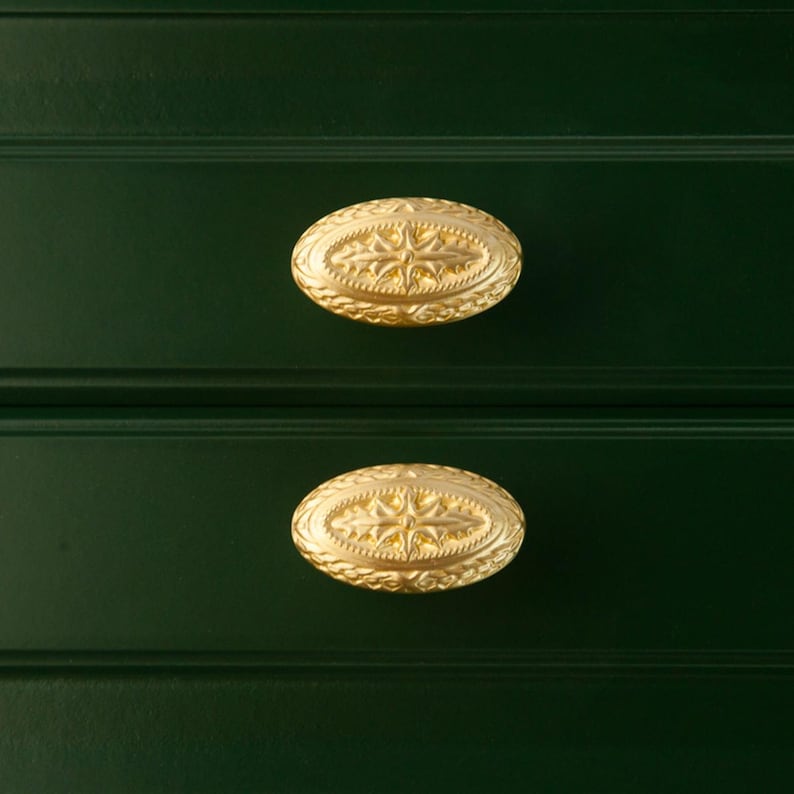 Puede incluir: Primer plano de dos tiradores de caj&oacute;n ovalados dorados con un dise&ntilde;o de estrella sobre un mueble verde oscuro. Los tiradores tienen una superficie detallada y texturizada.